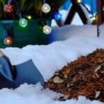 Zimný mulč a záhradná lopata na snehu s vianočnými svetlami a zasneženými rastlinami